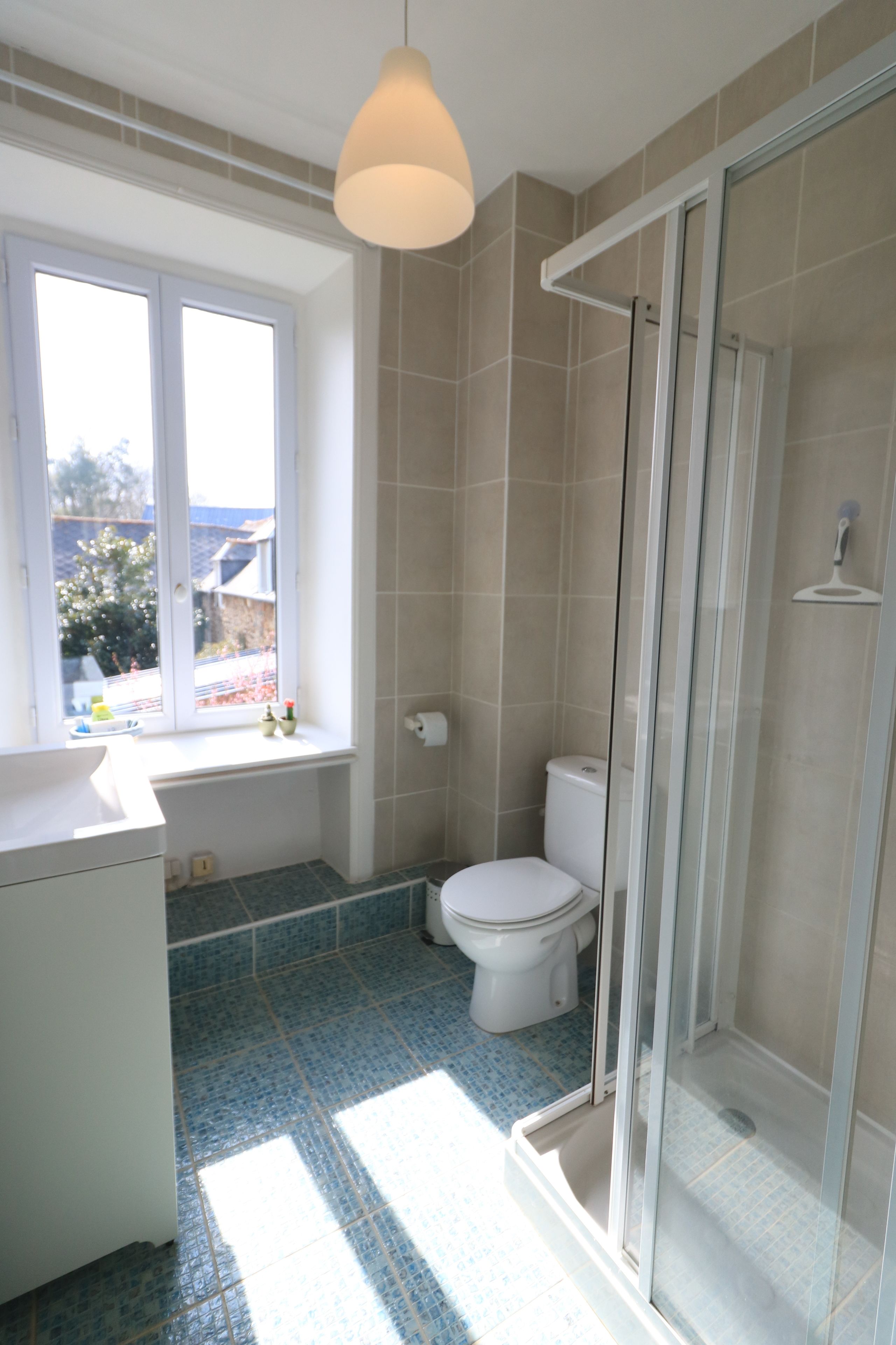 Un baño con inodoro, lavamanos, ducha con puerta de cristal y ventana que proporciona luz natural.