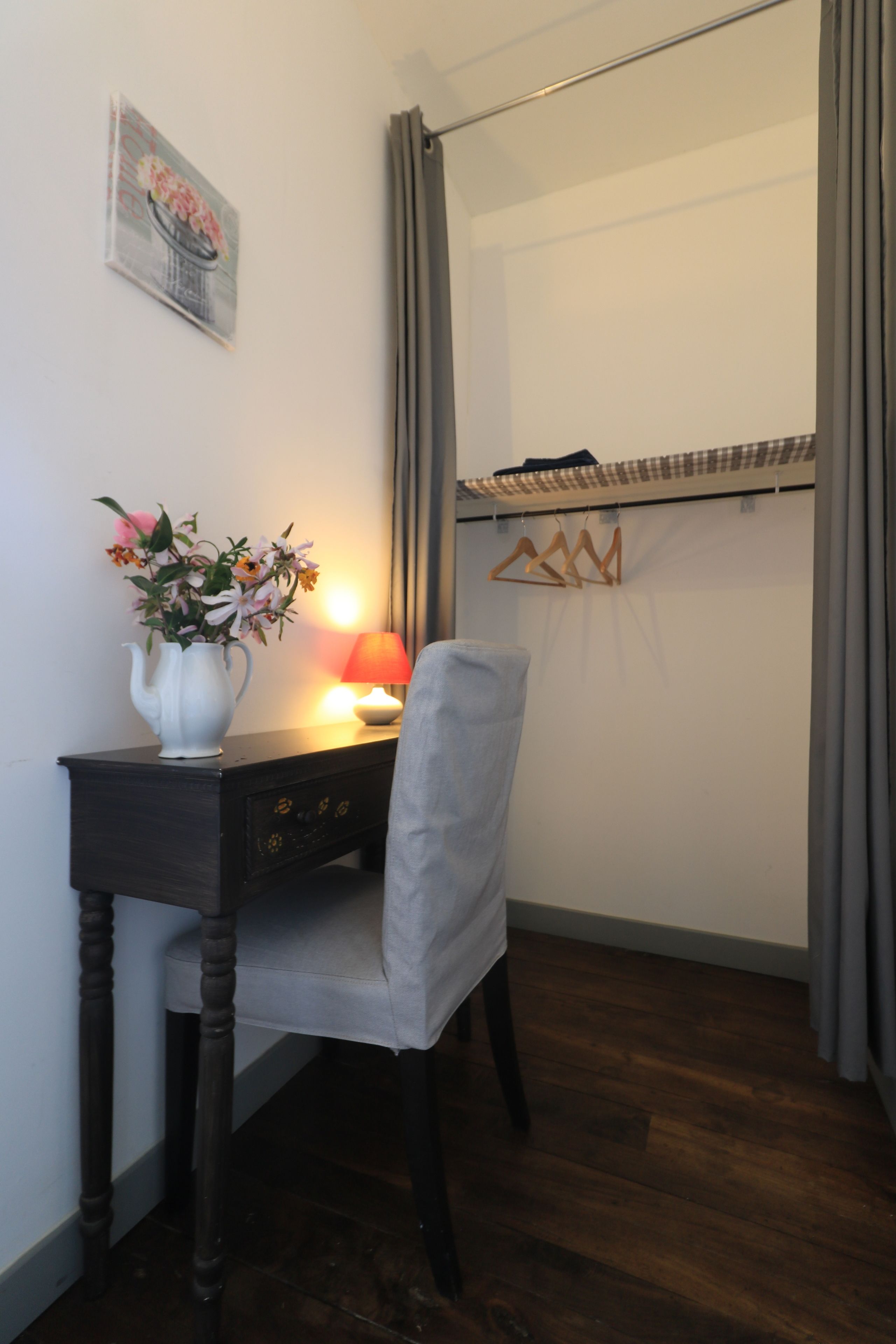 A small, neatly arranged bedroom with a desk, chair, and a bedside table with a lamp.