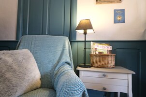 Una habitación con un sillón estampado, una mesita de noche con lámpara, una cesta y un libro titulado 'maison' en la pared.