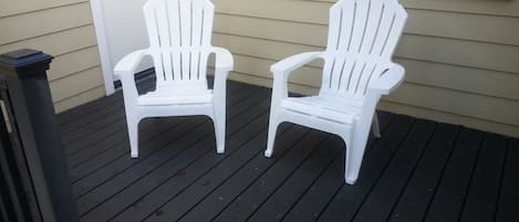 Two white Adirondack chairs on a wooden deck with a white door and window in the background.