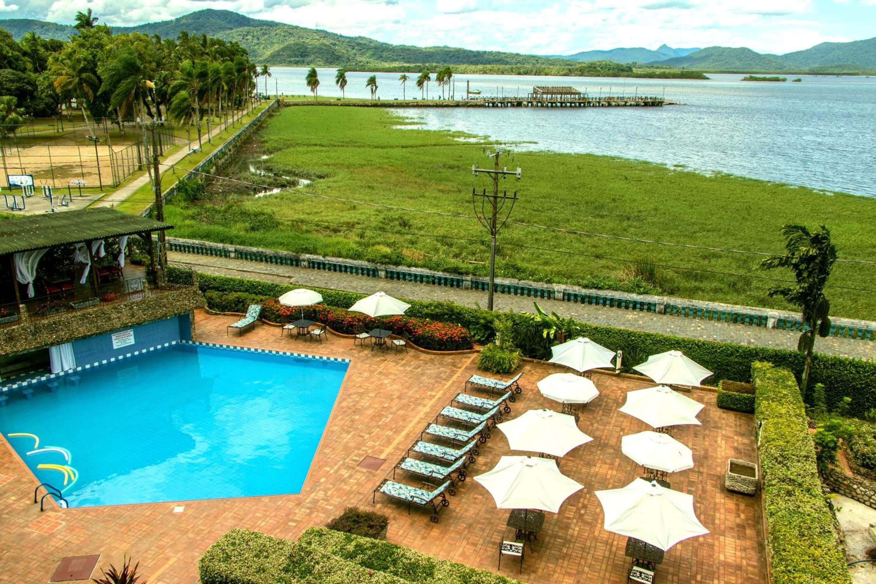 Piscina con sillas de descanso y sombrillas, rodeada de jardines y con vista a un lago y montañas.