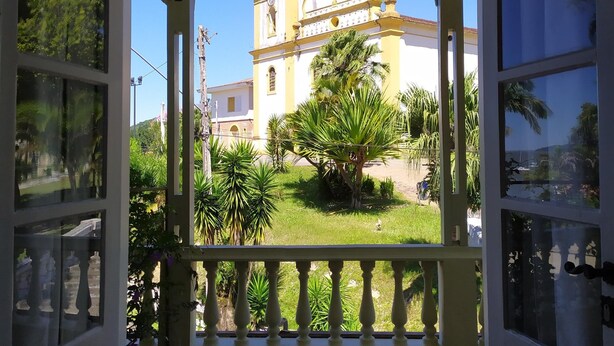 Un ventanal abierto que da a un balcón con vistas a una iglesia amarilla con campanario y una palmera en primer plano.