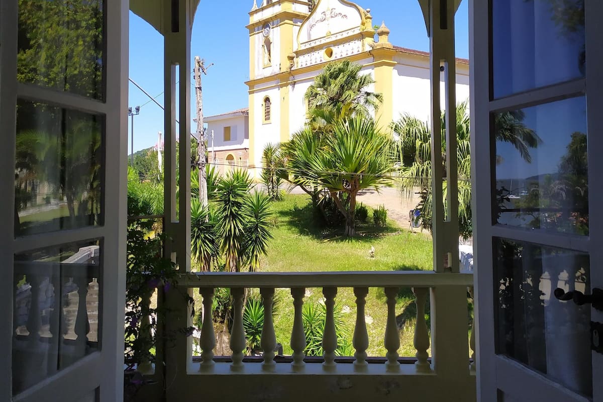 Eine geöffnete Tür, die zu einem Balkon mit Blick auf eine gelbe Kirche mit Glockenturm und einer im Vordergrund stehenden Palme führt.