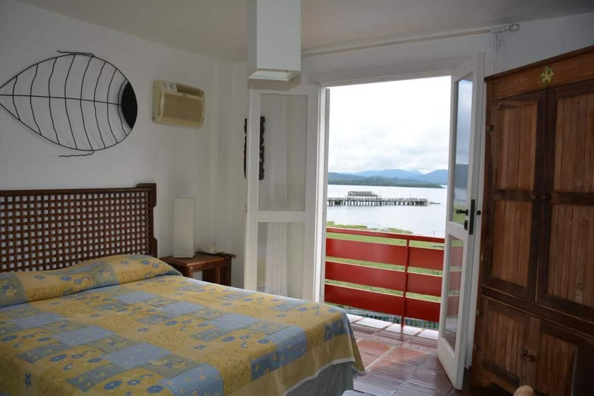 Ein Schlafzimmer mit Bett, Holz-Kopfteil, Wand-Klimaanlage und einer Tür zu einem Balkon mit Blick auf ein Gewässer und Berge.