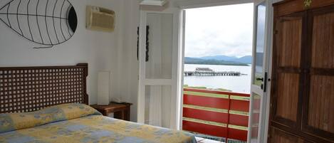 A bedroom with a bed, a wooden headboard, a wall-mounted air conditioner, a door leading to a balcony with a view of a body of water and mountains.