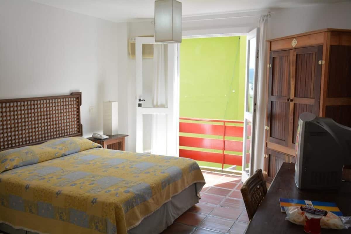 Ein Hotelzimmer mit Bett, Fernseher, einem Tisch mit Snacks und Blick auf ein rotes und grünes Gebäude.
