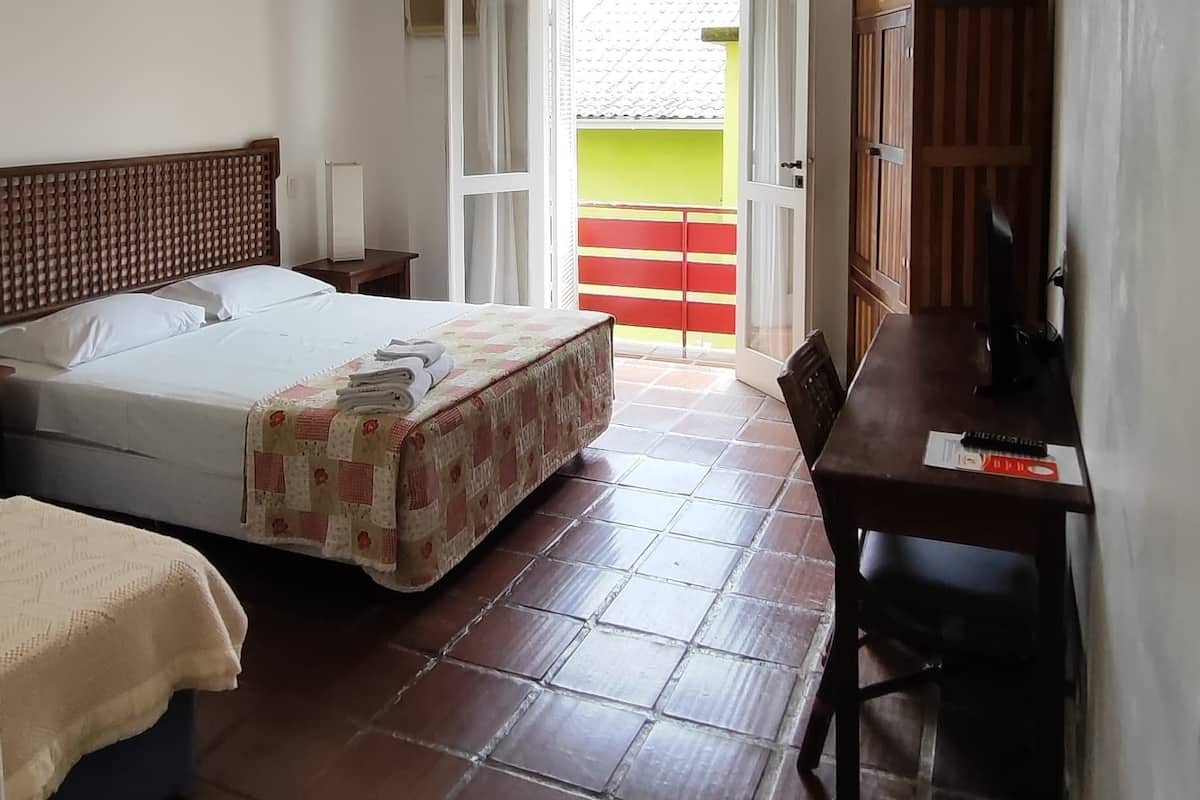 Ein Zimmer mit einem Bett, einem Schreibtisch und einem Stuhl. Ein Fenster bietet Blick auf einen Balkon.