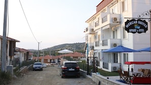 A street view with a Mercan Suites building, featuring balconies, outdoor seating, and a dirt road. - Mercan Suite Hotel (Gökçeada)