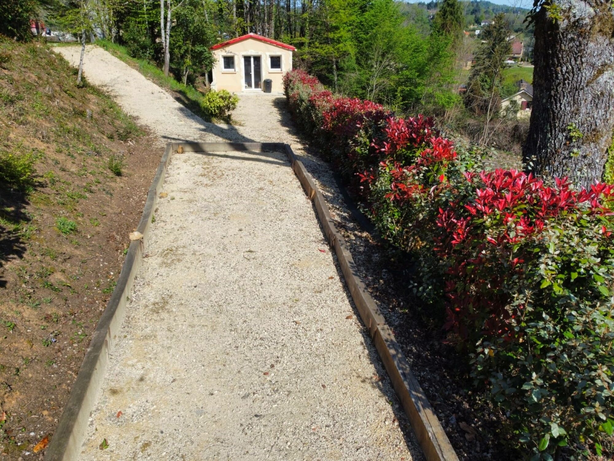 Uma trilha de cascalho leva a um pequeno edifício com telhado vermelho, cercado por vegetação e árvores.