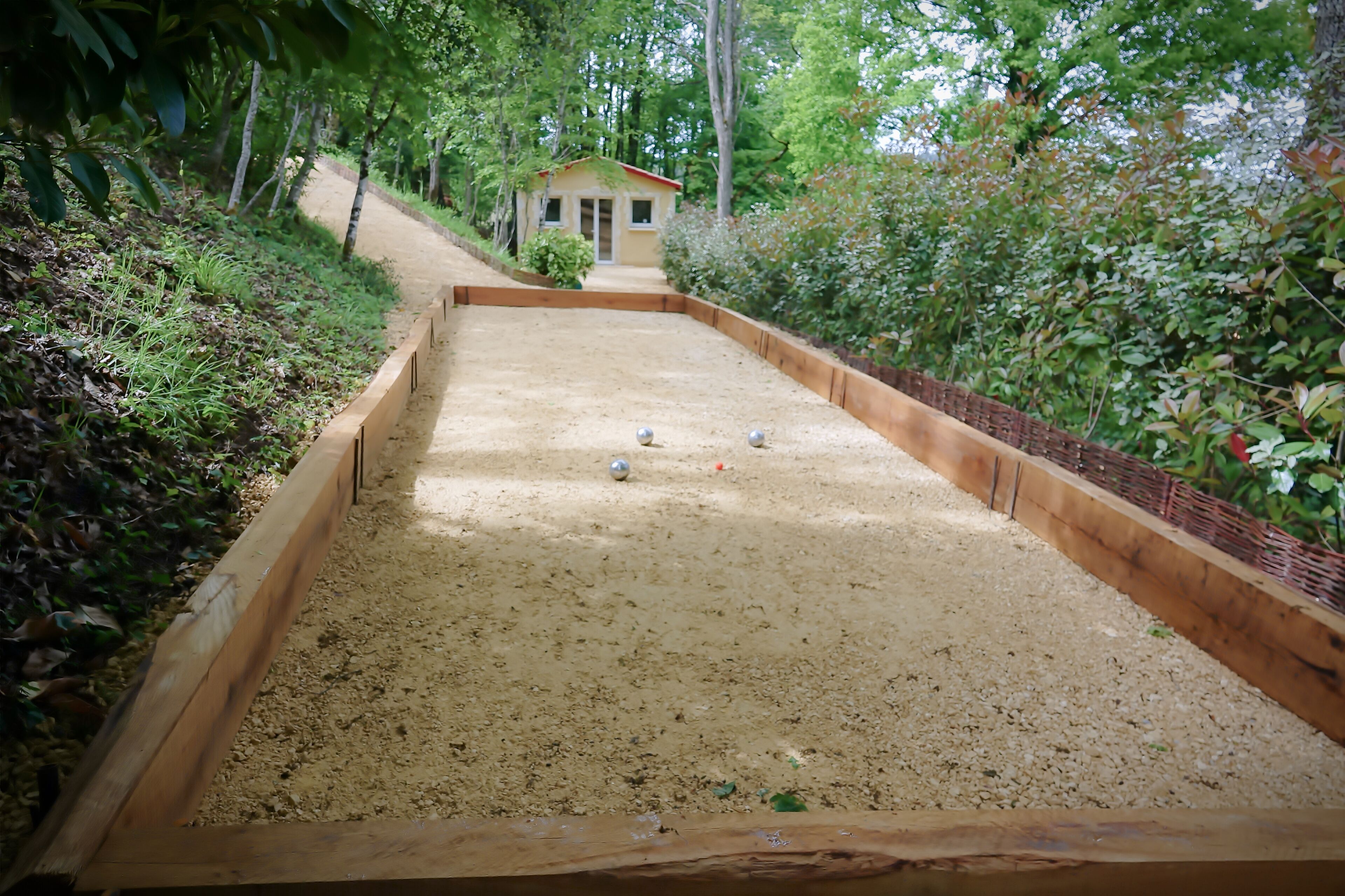 Une table de billard avec trois billes et un bord en bois, entourée de verdure et avec un petit bâtiment en arrière-plan.