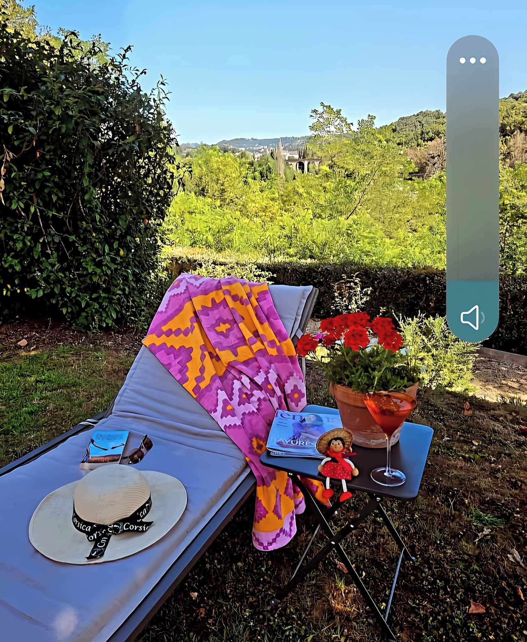 Una tumbona con una manta estampada, un sombrero, una revista, una figurita y una copa de martini sobre una mesa.