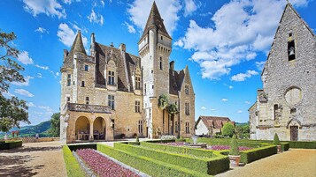 Un castillo histórico con una torre central, rodeado de jardines cuidados y un edificio más pequeño adyacente.