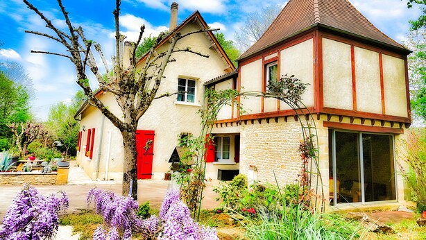 Una casa de dos pisos con puerta roja y fachada de piedra, rodeada de un jardín con flores moradas.