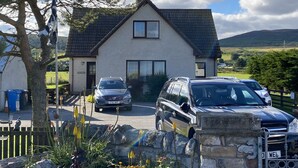 Uma casa de dois andares com telhado inclinado, entrada por carro e carros estacionados.
