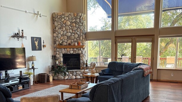 A spacious living room with a stone fireplace, large windows, and a ceiling fan. - Beautiful Mountain Home Near Yosemite (Groveland)