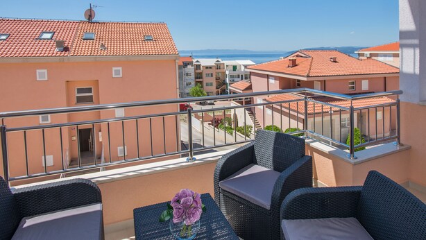 A balcony with wicker furniture and a laptop, overlooking a coastal area with terracotta-roofed buildings. - Apartman Dominik (Makarska)