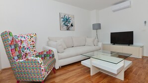 A living room with a colorful armchair, a white sofa, a glass coffee table, a flat-screen TV, and an air conditioner on the wall. - Apartman Dominik (Makarska)