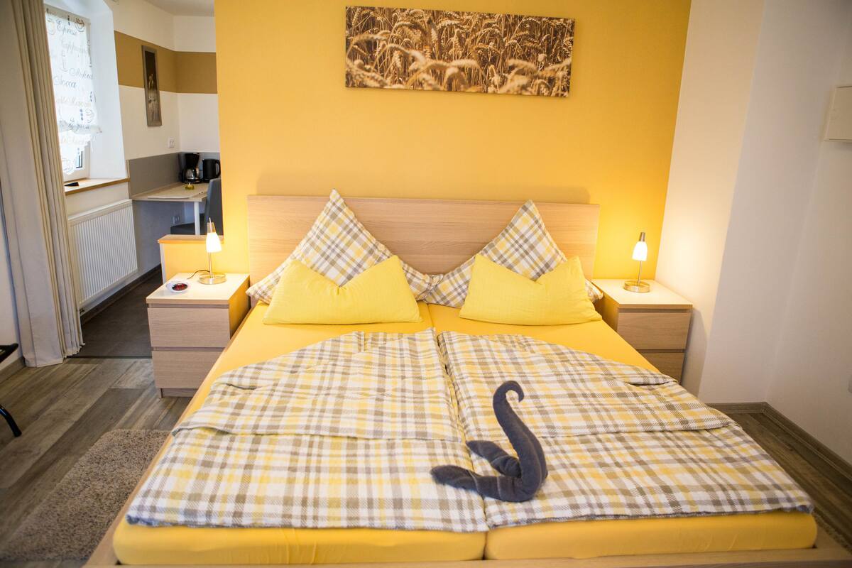 A hotel room with a checkered bedspread, two pillows, a bedside table with a lamp, and a kitchen area visible in the background.