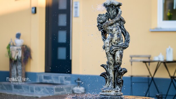 Eine Fontäne, aus deren Spitze Wasser sprüht, vor einer gelben Wand mit einer Tür und einem Fenster im Hintergrund.
