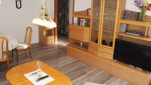 A living room with a wooden TV stand, a glass cabinet with books, a dining table with chairs, and a hanging lamp. - Ferienwohnung Braunlager Hexe (Braunlage)