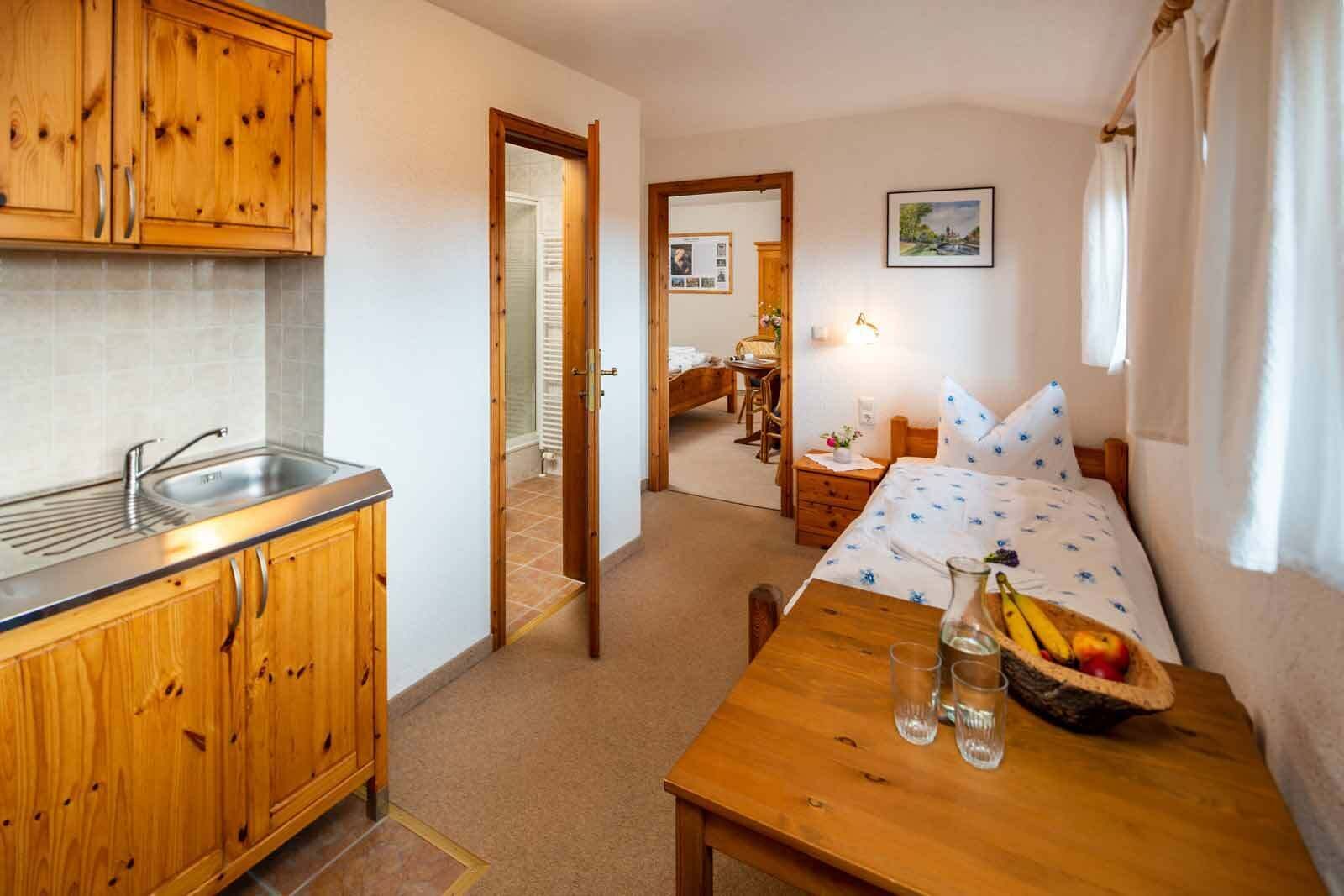 A compact room with wooden cabinets, a sink, a bed with a floral bedspread, a small table with fruit and glasses, and a desk with a chair.