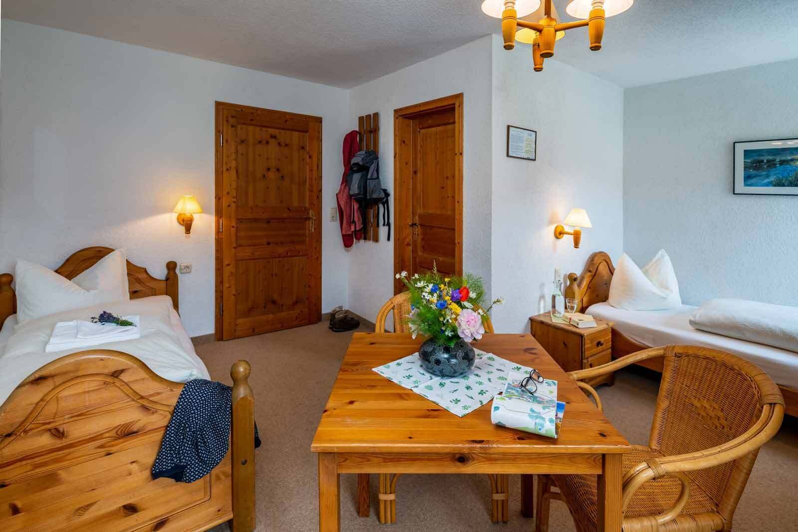 Habitación de hotel con dos camas de madera, una mesa de madera con un florero y una silla de mimbre.
