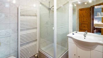 Un bany amb dutxa tancada amb vidre, un lavabo blanc i una porta de fusta.