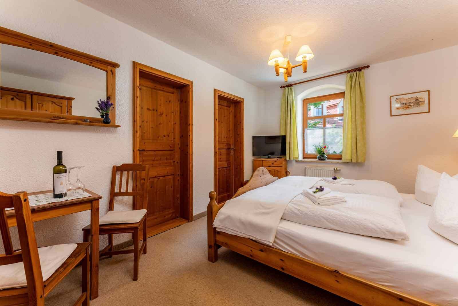 Un dormitorio con una cama de madera, una mesita de noche con una botella y vasos, una silla y un televisor sobre un aparador.