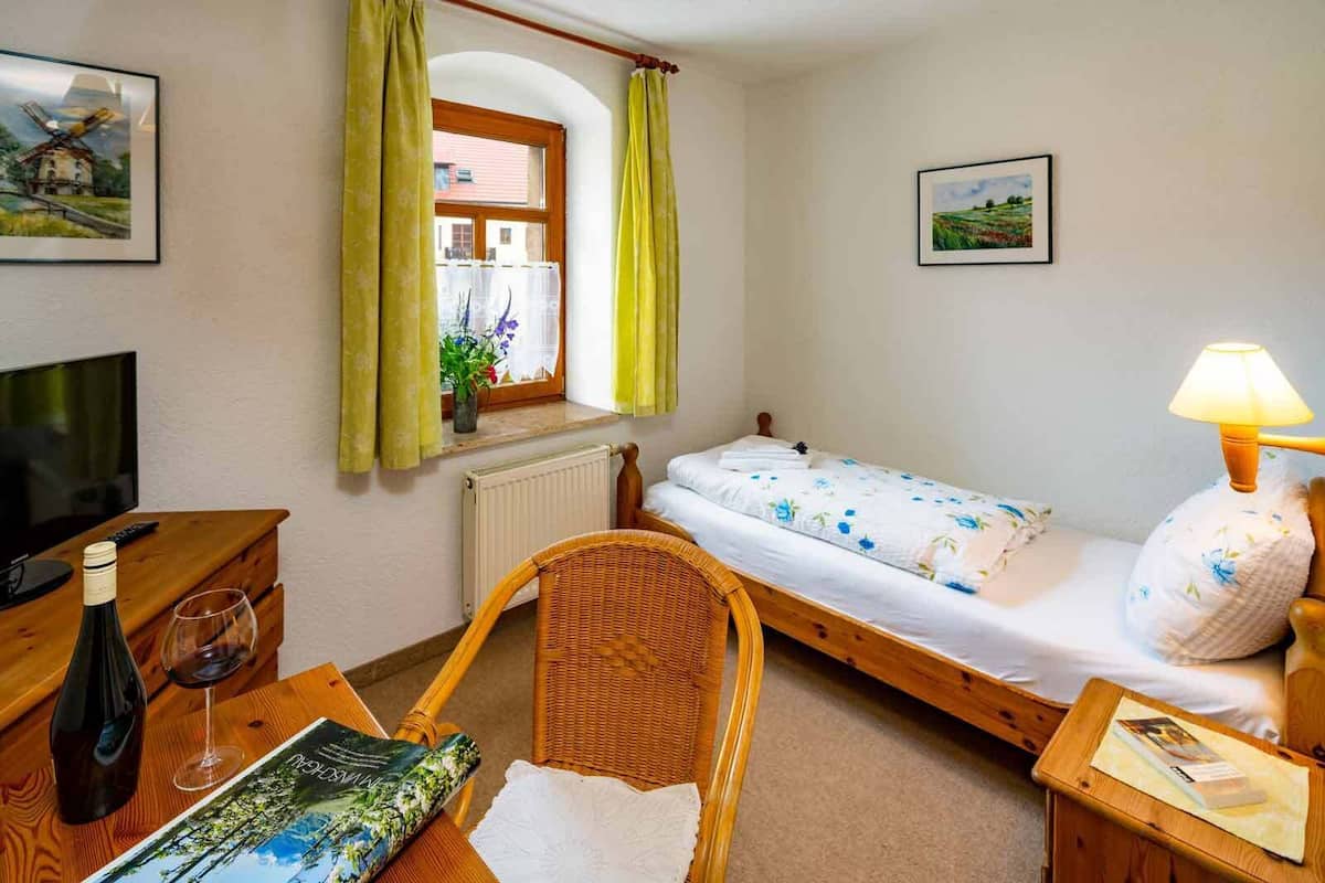 A cozy room with a single bed, a small table with a bottle and glass, a chair, a lamp, and two framed pictures on the walls.