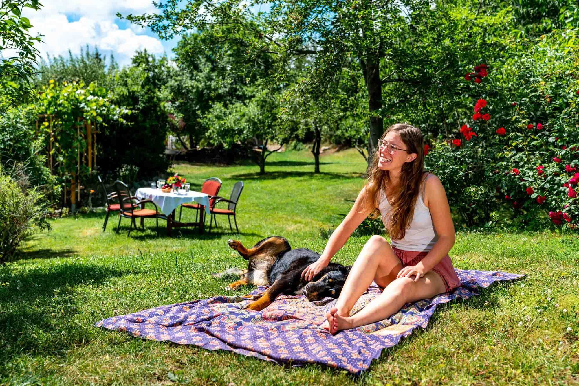 Una mujer sentada sobre una manta en un jardín, con un perro.
