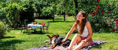 Una mujer sentada sobre una manta en un jardín, con un perro.