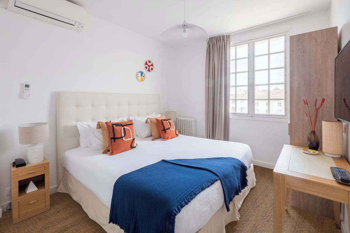 Une chambre d’hôtel avec un grand lit, un bureau, une chaise, une lampe, un radiateur, une fenêtre avec des rideaux et un climatiseur fixé au mur.