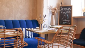 A cozy seating area with a blue sofa, wicker chairs, and a table with a glass pitcher and a vase of dried flowers. A chalkboard sign reads "Bienvenue au petit-déjeuner de la Tour de Nesle." - Hôtel La Tour de Nesle La Rochelle Vieux Port (La Rochelle)