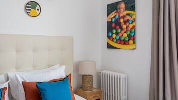 A neatly arranged bedroom with a bed, pillows, a bedside table with a lamp, a radiator, and two wall art pieces.