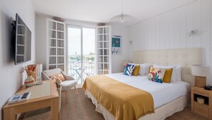 A hotel room with a bed, a desk, a chair, a TV, and a view of the city through large windows. - Hôtel La Tour de Nesle La Rochelle Vieux Port (La Rochelle)