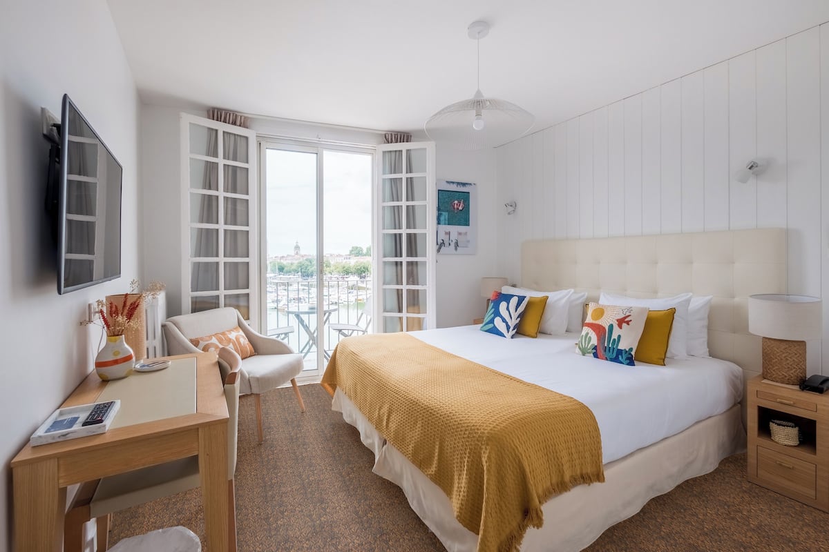 Une chambre d’hôtel équipée d’un lit, d’un bureau, d’une chaise, d’une télévision et offrant une vue sur la ville grâce à de grandes fenêtres.