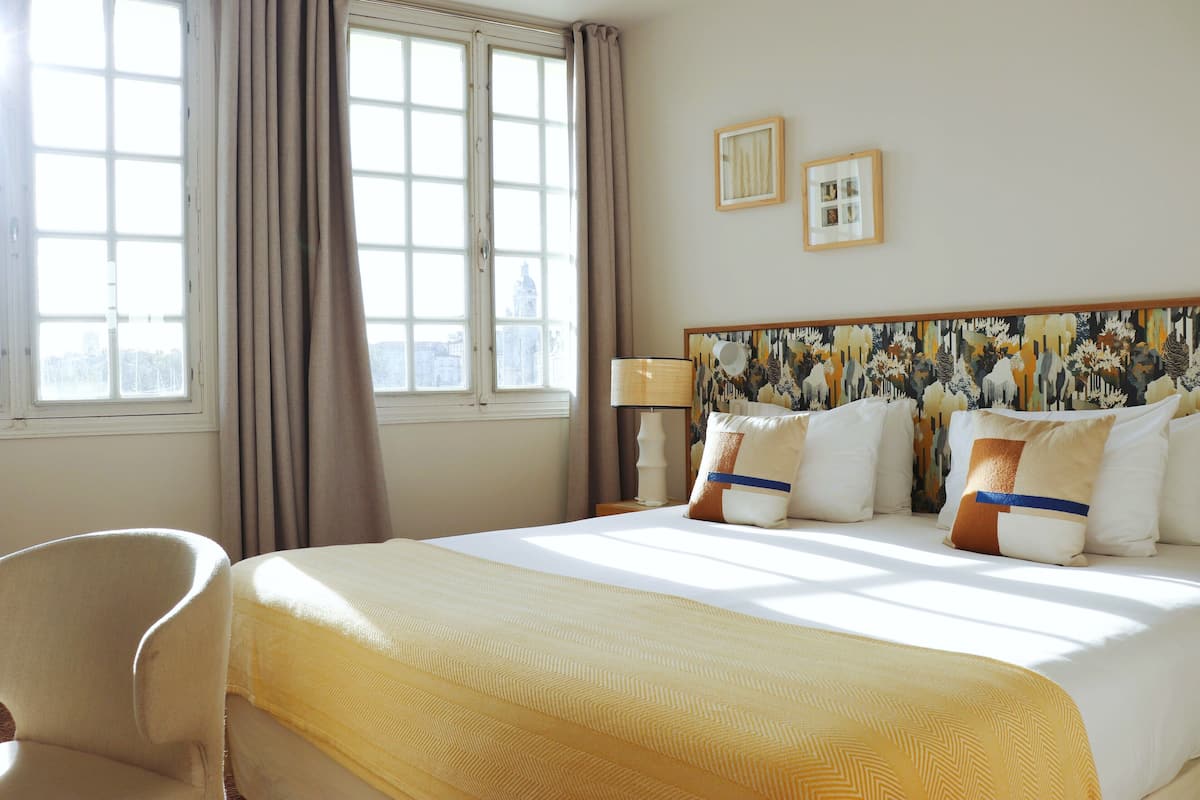 Une chambre d’hôtel avec un grand lit, une chaise, une fenêtre avec des rideaux et un tableau au mur.