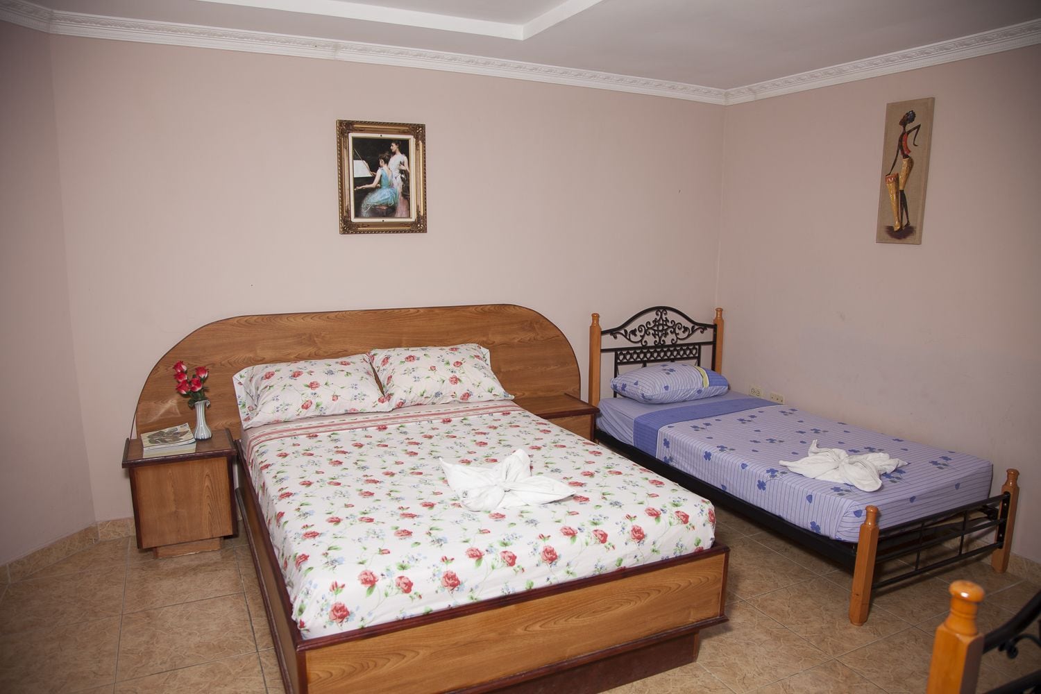 Un dormitorio con una cama de madera grande, una cama más pequeña, una mesita de noche con un florero y dos cuadros en las paredes.
