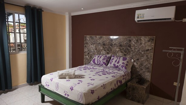 Un dormitorio con cama, mesita de noche, ventana con cortinas y un equipo de aire acondicionado.