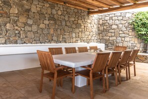 Un comedor al aire libre con un muro de piedra de fondo, sillas de madera y una mesa blanca.