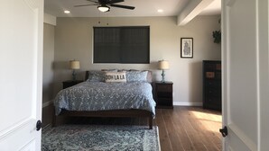 A bedroom with a bed, two bedside tables, a ceiling fan, a large window, and a dark wooden dresser. - Casa Tranquila Puerto Nuevo with Ocean and Mountain View that Sleeps 20 (Puerto Nuevo)