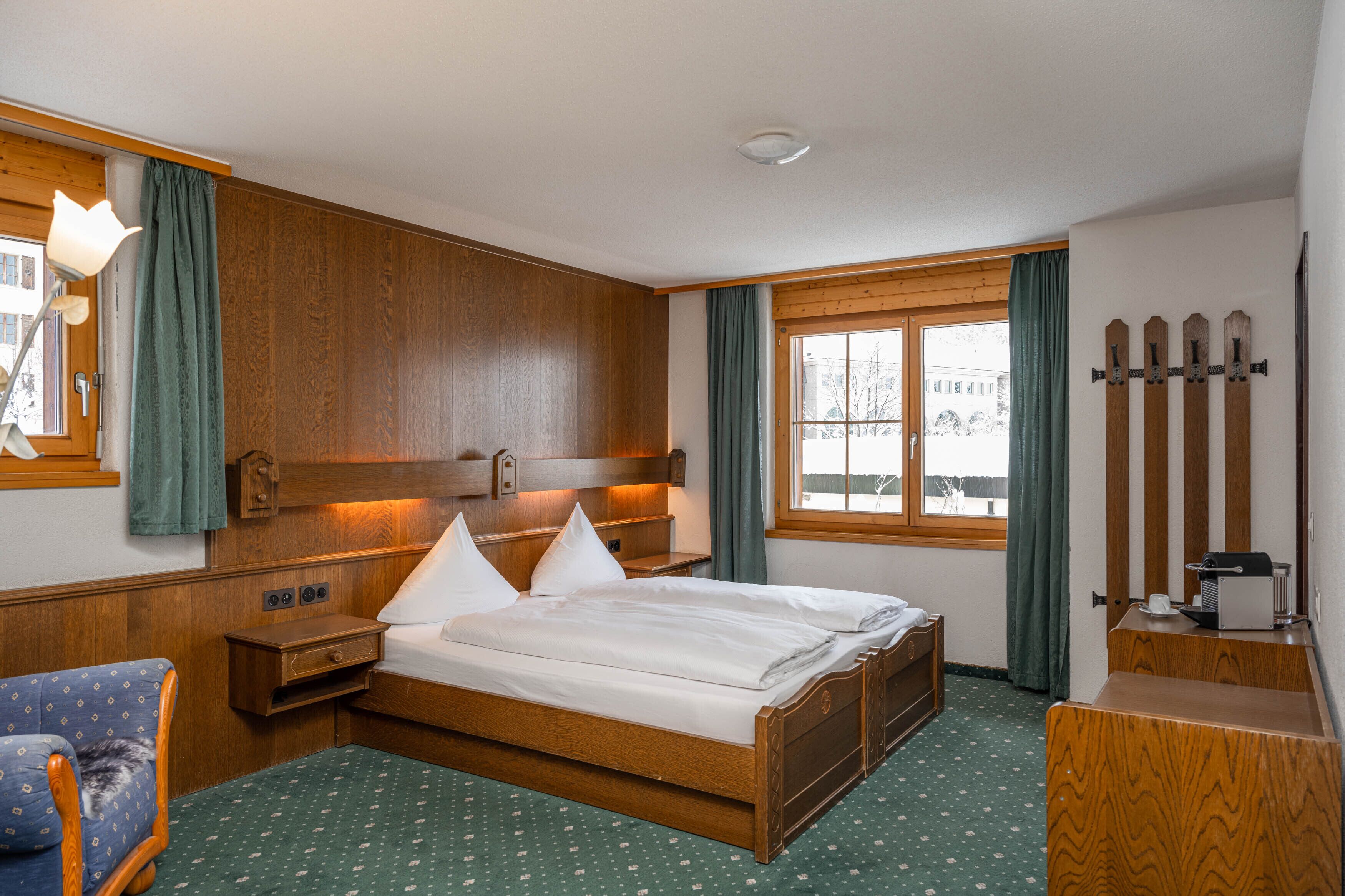 Habitación de hotel con una cama de madera, una mesita de noche pequeña, una silla, un escritorio y una ventana con cortinas.