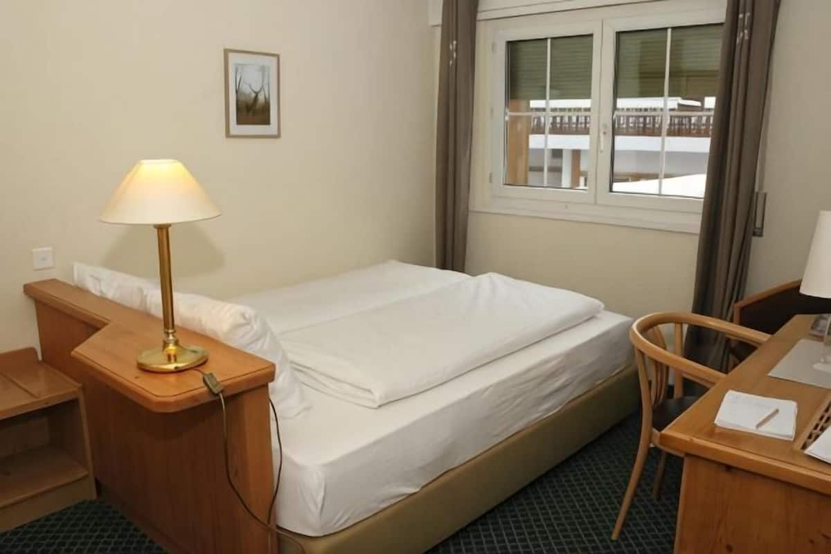 Habitación de hotel con cama, mesita de noche, lámpara, silla, escritorio y ventana.