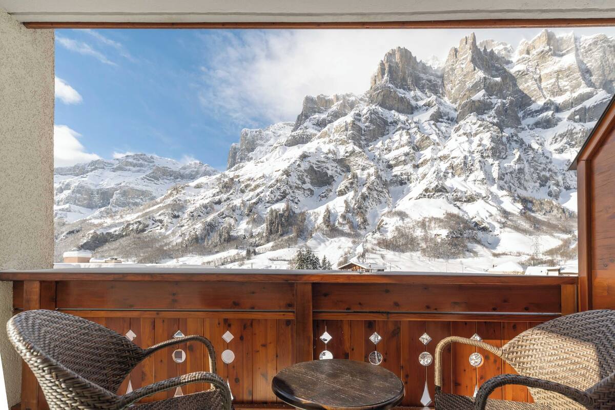 Un balcón de madera con sillas de mimbre y una mesa, con vistas a un paisaje montañoso nevado.