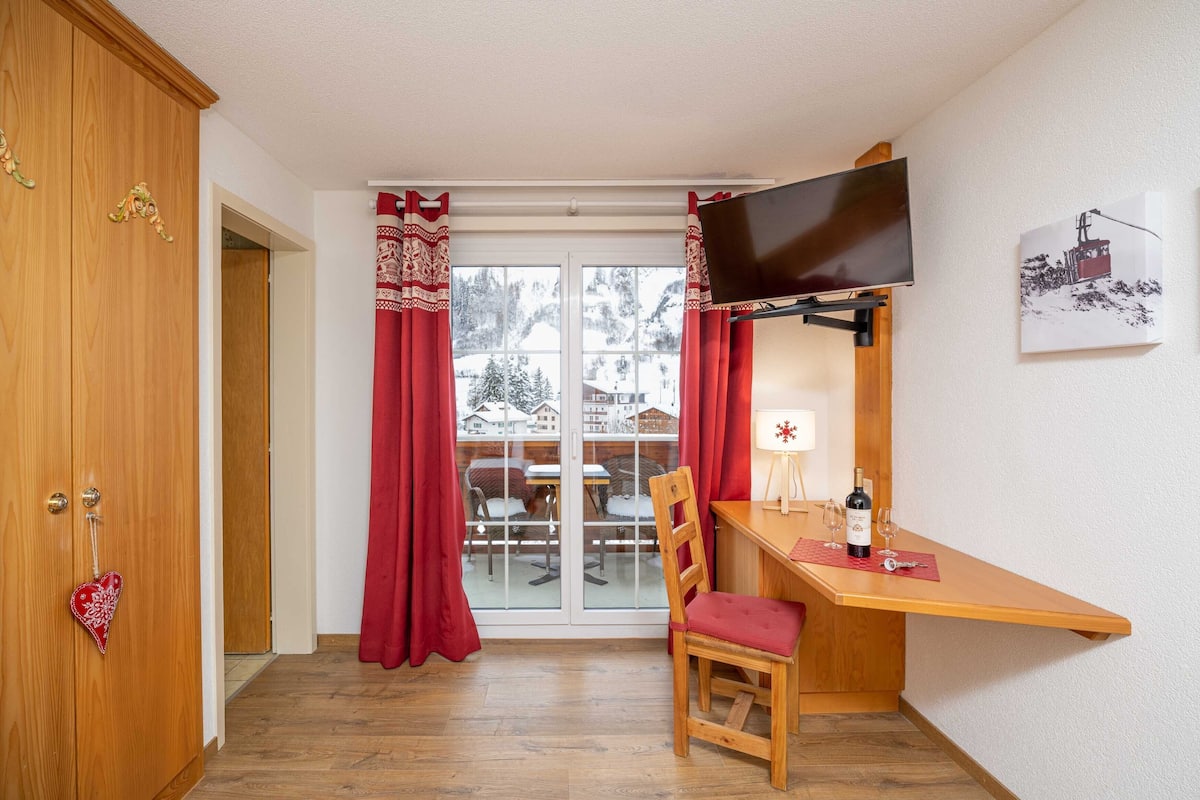 Una habitación con un escritorio de madera, una silla, un televisor montado en la pared y una ventana con cortinas rojas.