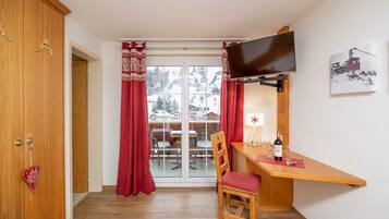 A room with a wooden desk, a chair, a TV mounted on the wall, and a window with red curtains.