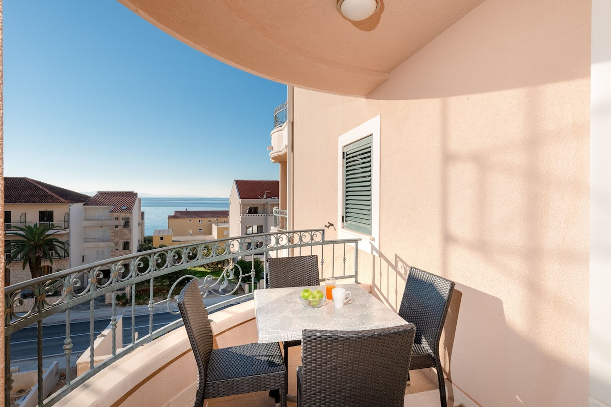 Ein Balkon mit Korbstühlen, einem Tisch mit einer Obstschale und einem Glas Orangensaft, und Blick auf das Meer und Gebäude.
