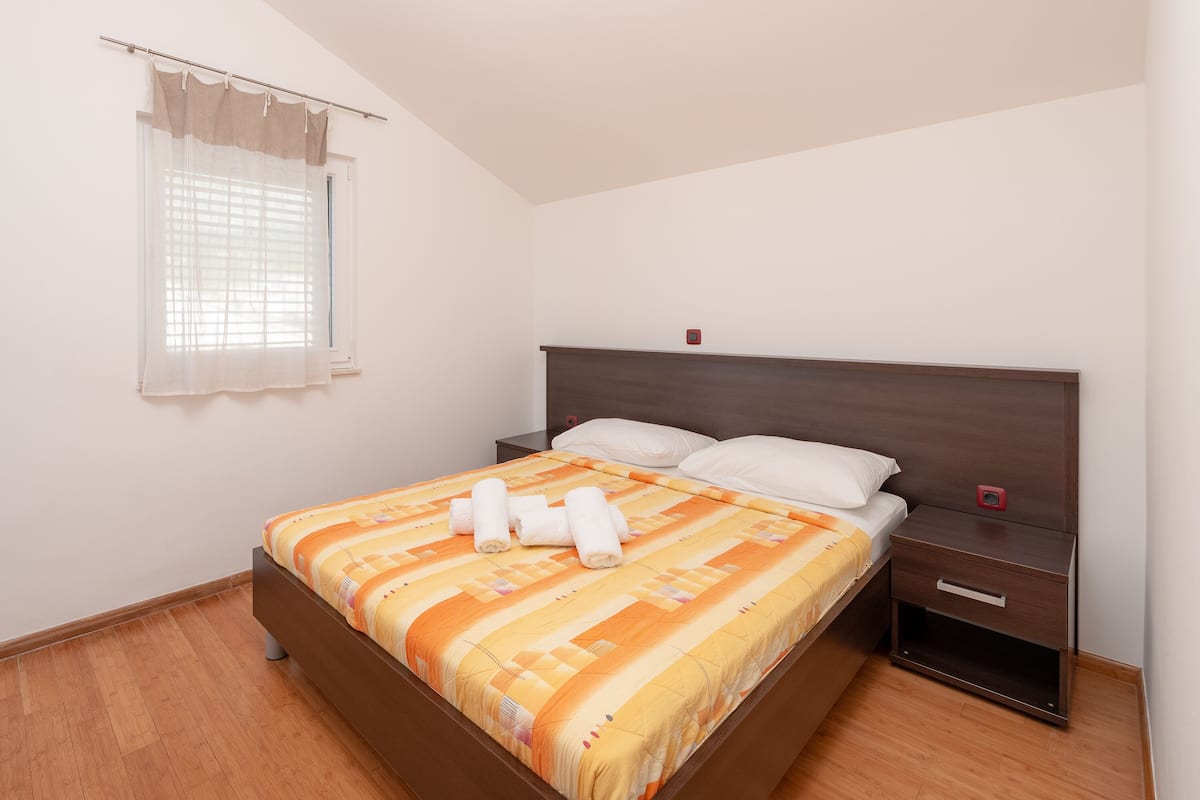 Ein ordentlich bezogenes Bett mit einem gemusterten Überwurf, ein Nachttisch mit Schublade und ein Fenster mit Jalousien.