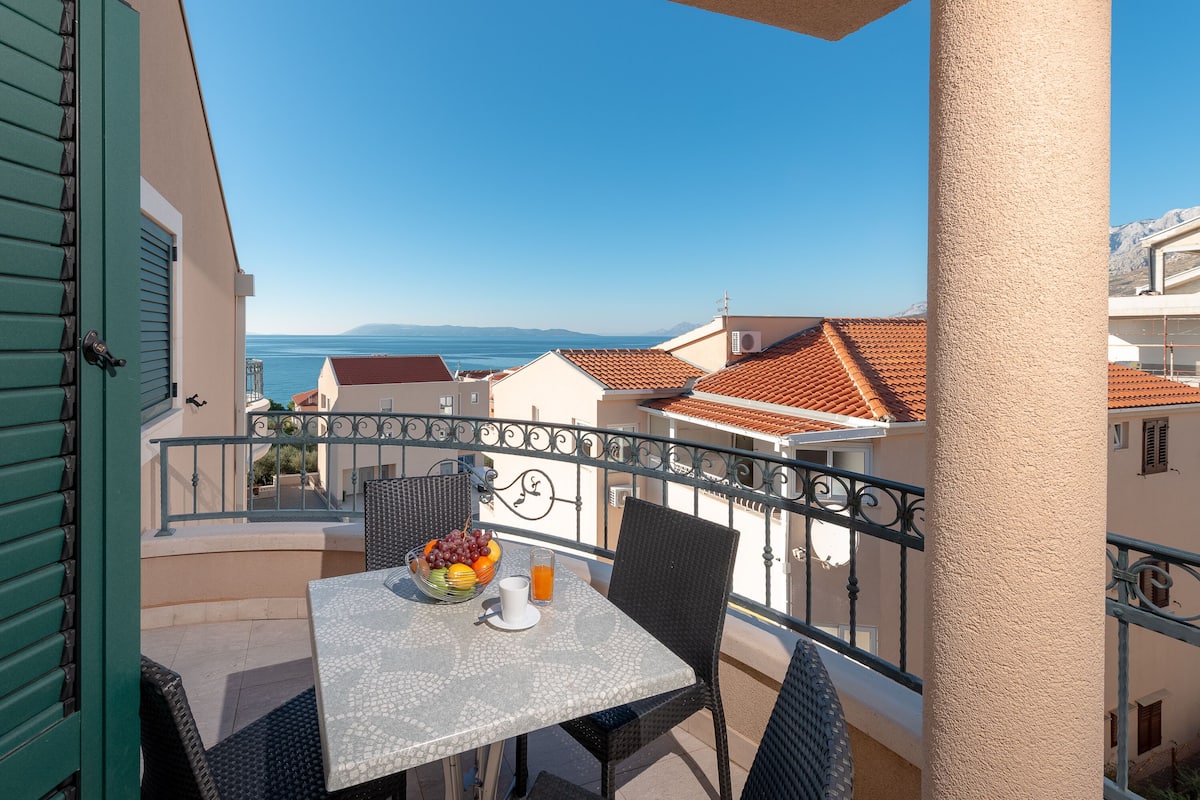 Ein Balkon mit Tisch für zwei Personen, einer Obstschale und einem Glas Saft. Der Balkon bietet Blick auf eine Küstenstadt mit Häusern mit Terrakotta-Dächern und blauem Himmel.