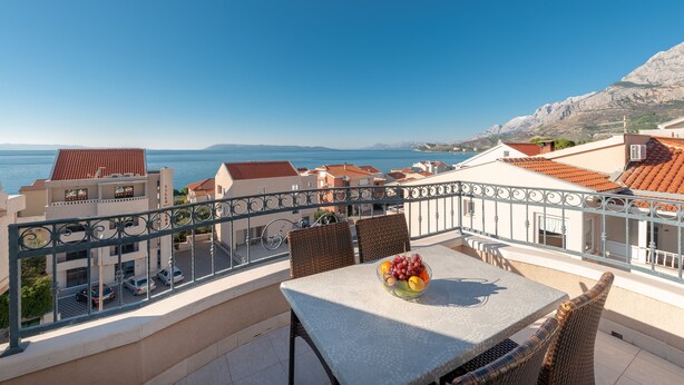 Un balcó amb taula i cadires, un plat de fruita i vista al mar i edificis.
